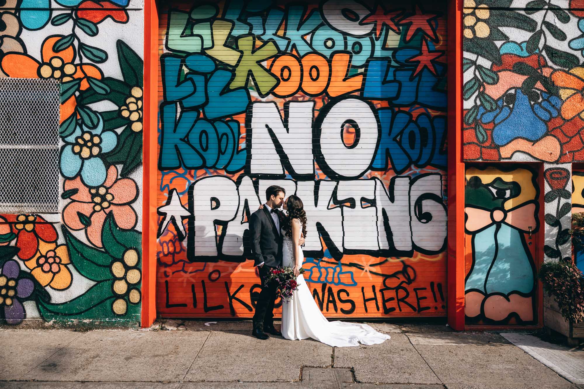 Bride and groom standing in front of a colorful floral wall full of street art. Brooklyn NY